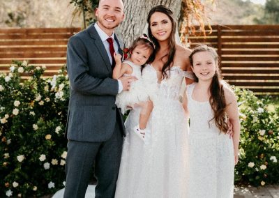 Bride and groom with two young girls beneath a grand oak framed by white blooms