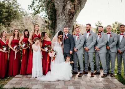 Wedding party in red dresses and gray suits with the couple beneath a large oak