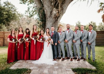 Full wedding party in red dresses and gray suits gathered under a sprawling oak