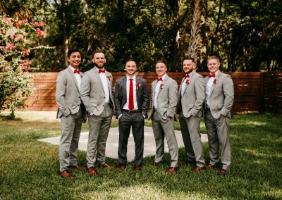 Groom in a charcoal suit and red tie with five groomsmen in gray suits on the lawn