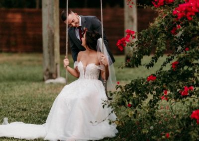 Groom leaning toward his bride seated on a garden swing near red bougainvillea