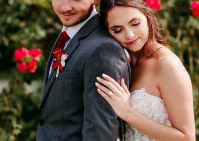 Bride resting her head on the groom's shoulder in front of bright red rose bushes