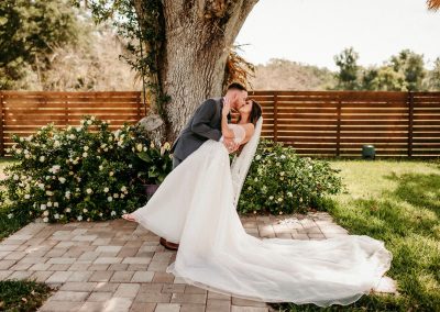Groom dipping his bride for a kiss under a grand oak, her gown train trailing across brick pavers