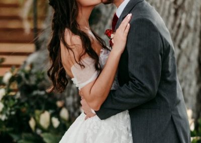 Bride and groom kissing beside a large oak, groom in a gray suit with a red boutonniere