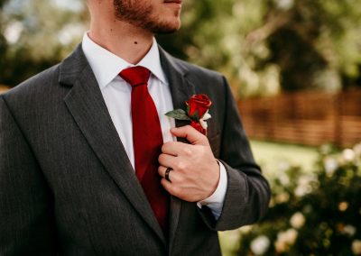 Groom in a gray suit and red tie adjusting his red rose boutonniere