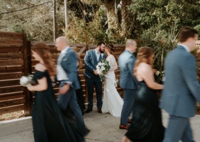 Wedding party in motion around a bride and groom sharing a kiss by a wood fence