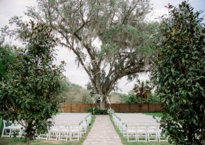Outdoor ceremony setup with white chairs and an aisle beneath a moss-draped oak