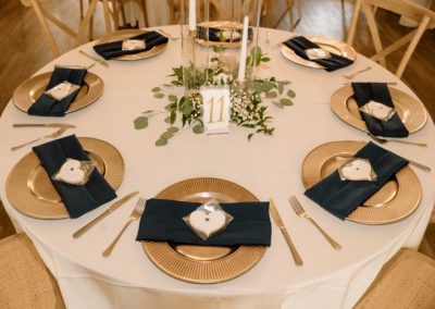 Round reception table with gold chargers, navy napkins, and a greenery centerpiece at table eleven