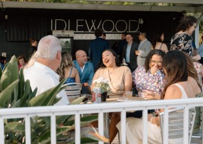 Wedding guests laughing around an outdoor table beneath the Idlewood sign