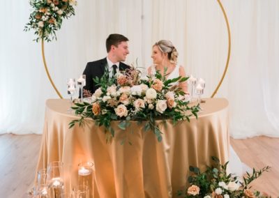 Couple at a gold sweetheart table framed by a round floral arch and candles
