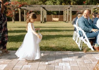 Flower girl in a white dress scattering petals down the paver aisle as guests watch