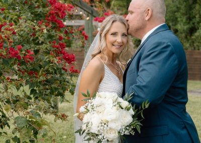 Groom kissing bride's forehead beside red bougainvillea as she holds a white bouquet