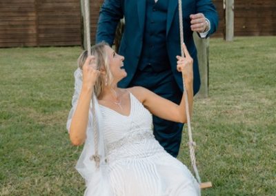 Bride on a rope swing laughing up at her groom in a navy suit on the lawn