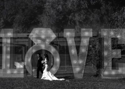 Bride and groom walking past a glowing LOVE marquee at night, black and white