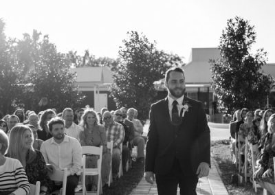 Groom walking down the outdoor aisle between seated guests, in black and white