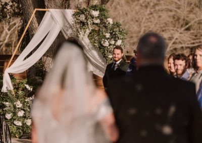 Groom watching his bride approach beneath a draped hexagon arch and white florals