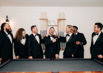Groomsmen in black tuxedos laughing together beside a wall of pool cues