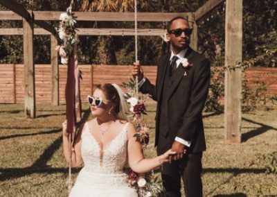 Bride in heart sunglasses on a floral swing with the groom in a black suit beside her