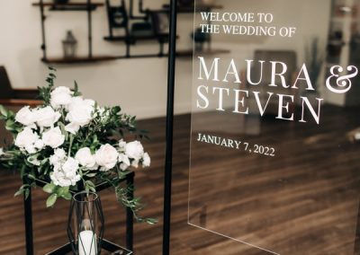 Acrylic welcome sign for Maura and Steven beside a white rose arrangement
