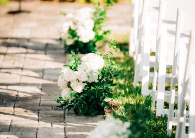 White floral arrangements lining a paver aisle beside rows of white folding chairs