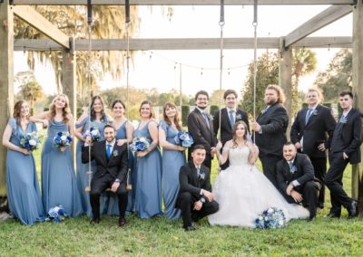 Wedding party in dusty-blue gowns and navy suits around a bride on a swing at sunset