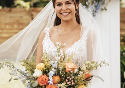 Bride in a veil holding a vibrant orange and blue bouquet before a floral arch