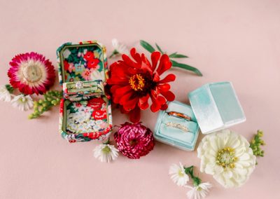 Wedding rings in velvet boxes surrounded by bright zinnias on a pink backdrop