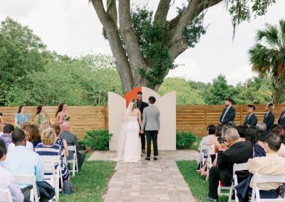 Outdoor ceremony beneath a grand oak, the couple at an arched backdrop before seated guests