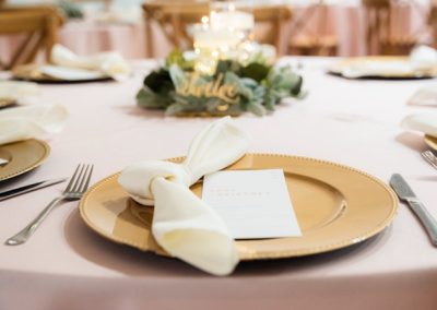 Gold charger plate with a knotted ivory napkin and menu card on a blush-linen reception table