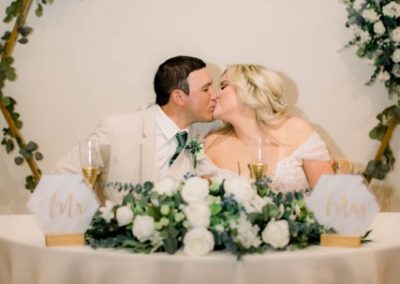 Newlyweds kissing at their sweetheart table under a hexagon greenery arch with a C monogram