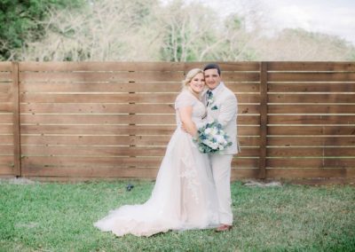 Smiling bride and groom posing against a wood-plank fence with a blue-and-white bouquet