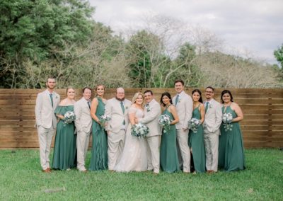 Wedding party in sage green gowns and tan suits on the lawn by a wood fence