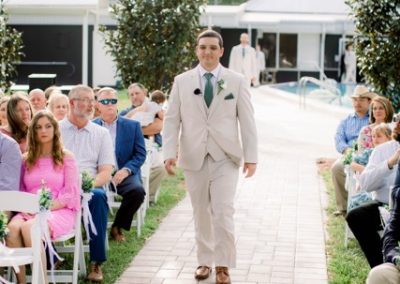 Groom walking down the paver aisle between seated guests at a poolside ceremony