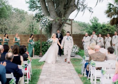 Newlyweds recess down the aisle as their wedding party cheers beneath the oak