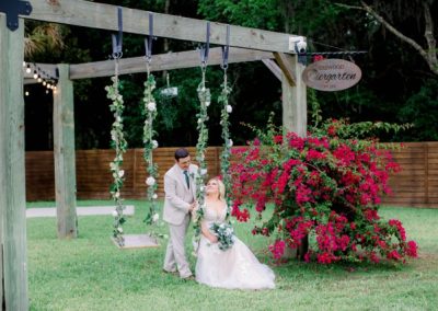 Groom and bride on a greenery-draped swing beside bougainvillea at the Biergarten pergola