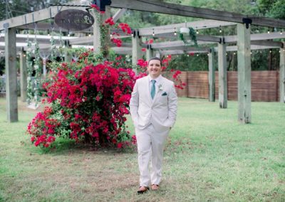 Groom in a tan suit standing beside bright pink bougainvillea under the pergola