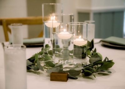 Floating candles in glass cylinders ringed with eucalyptus on a white reception table