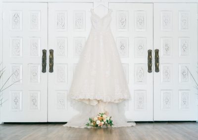 Lace wedding gown hanging against ornate carved white doors above a bouquet
