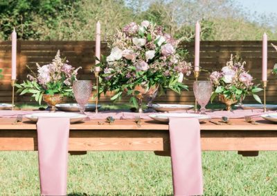 Outdoor farm table set with mauve florals, dusty rose napkins and gold candlesticks on the lawn
