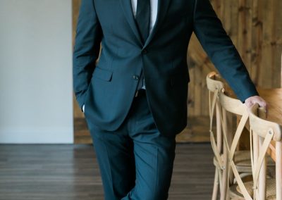 Groom in a dark suit smiling beside a rustic barn door and crossback chairs