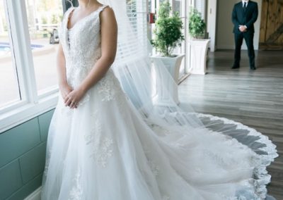 Bride in a lace gown by tall windows as the groom waits behind for the first look