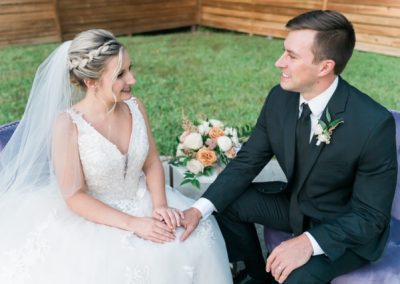 Bride and groom holding hands on a velvet loveseat outdoors beside a floral arrangement