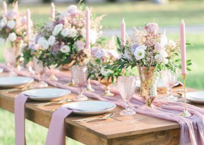 Wooden farm table dressed with blush blooms, pink goblets and taper candles outdoors